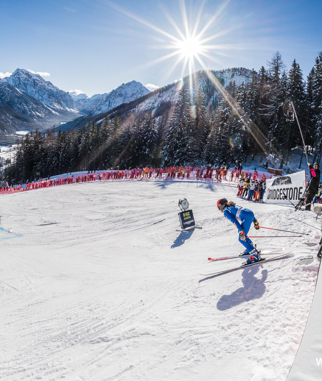 Eine Skirennläuferin startet den Riesenslalom und wird von Zuschauern am Rande der Piste bejubelt, im Hintergrund das Bergpanorama und strahlend blauer Himmel. | © Harald Wisthaler