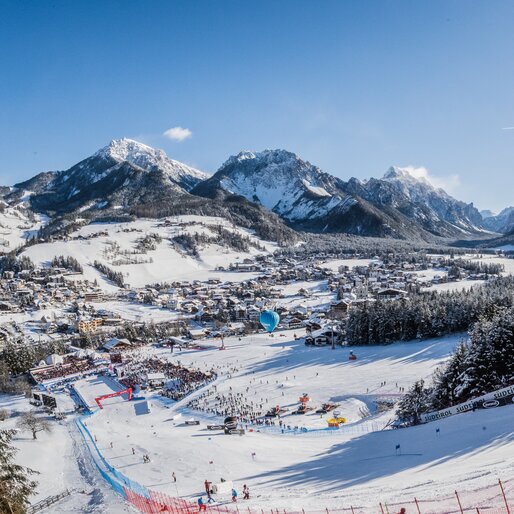 Skirennstrecke und Ortschaft im Vordergrund und blauer Himmel, verschneite Wälder und Berge im Hintergrund. | © Harald Wisthaler