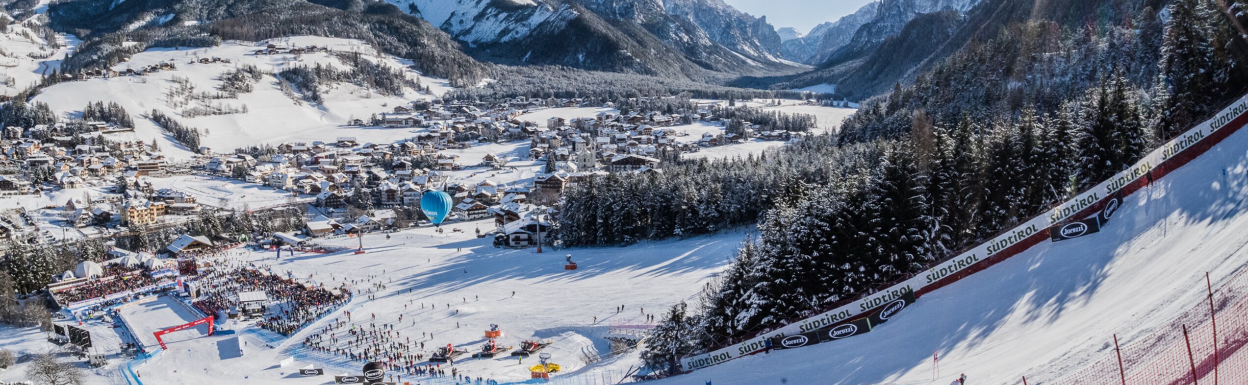 Skirennstrecke und Ortschaft im Vordergrund und blauer Himmel, verschneite Wälder und Berge im Hintergrund. | © Harald Wisthaler