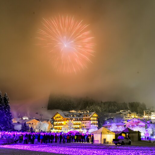 Feuerwerk nachts über einem verschneiten Skiort, im Vordergrund eine lila beleuchtete Bühne mit Zuschauern. | © Gianvito Coco