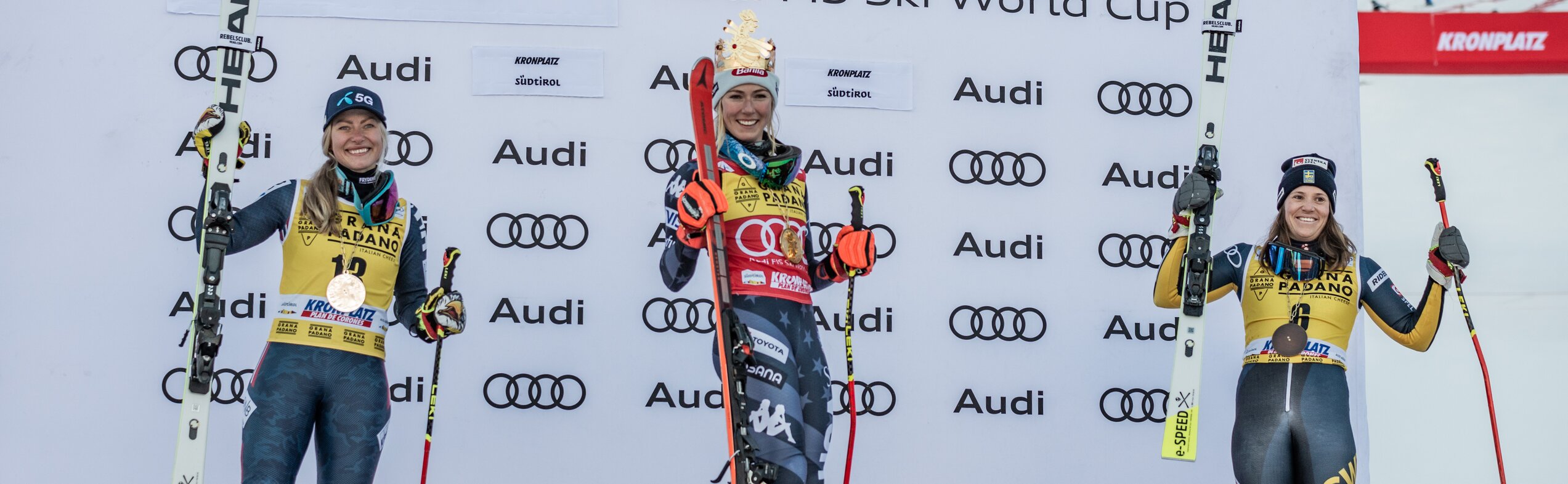 Drei jubelnde Skirennläuferinnen auf dem Podest, die mittlere trägt eine Krone. | © Gianvito Coco