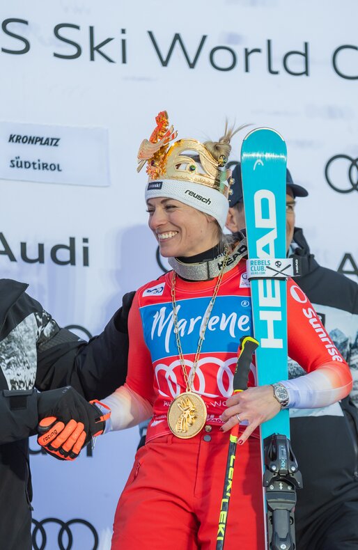 Eine Skirennläuferin im roten Rennanzug steht auf dem Siegerpodest und trägt eine goldene Krone, während sie einem Mann die Hand schüttelt. | © Gianvito Coco