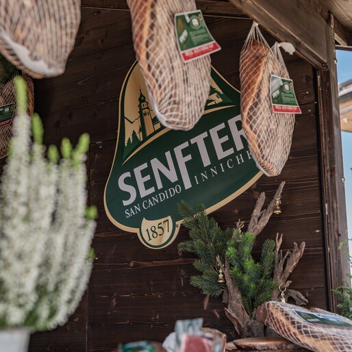Ein Stand mit Senfter-Produkten zeigt hängende Speckstücke und regionale Spezialitäten unter einem Vordach. | © Gianvito Coco