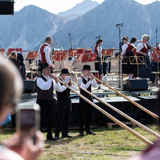Drei Männer stehen in traditioneller Tracht auf einer Bergwiese und spielen Alphorn, im Hintergrund die Berge und eine Musikkapelle.