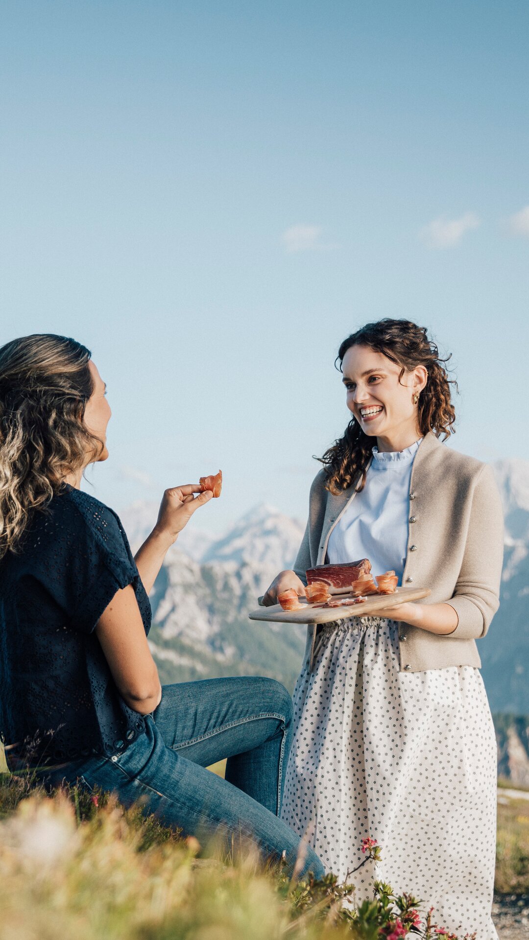 Vor dem Bergpanorama sieht man eine stehende Frau mit einem Brett Speck in den Händen und sie lächelt eine sitzende Frau an, welche sich an dem Speck bedient. | © IDM Südtirol