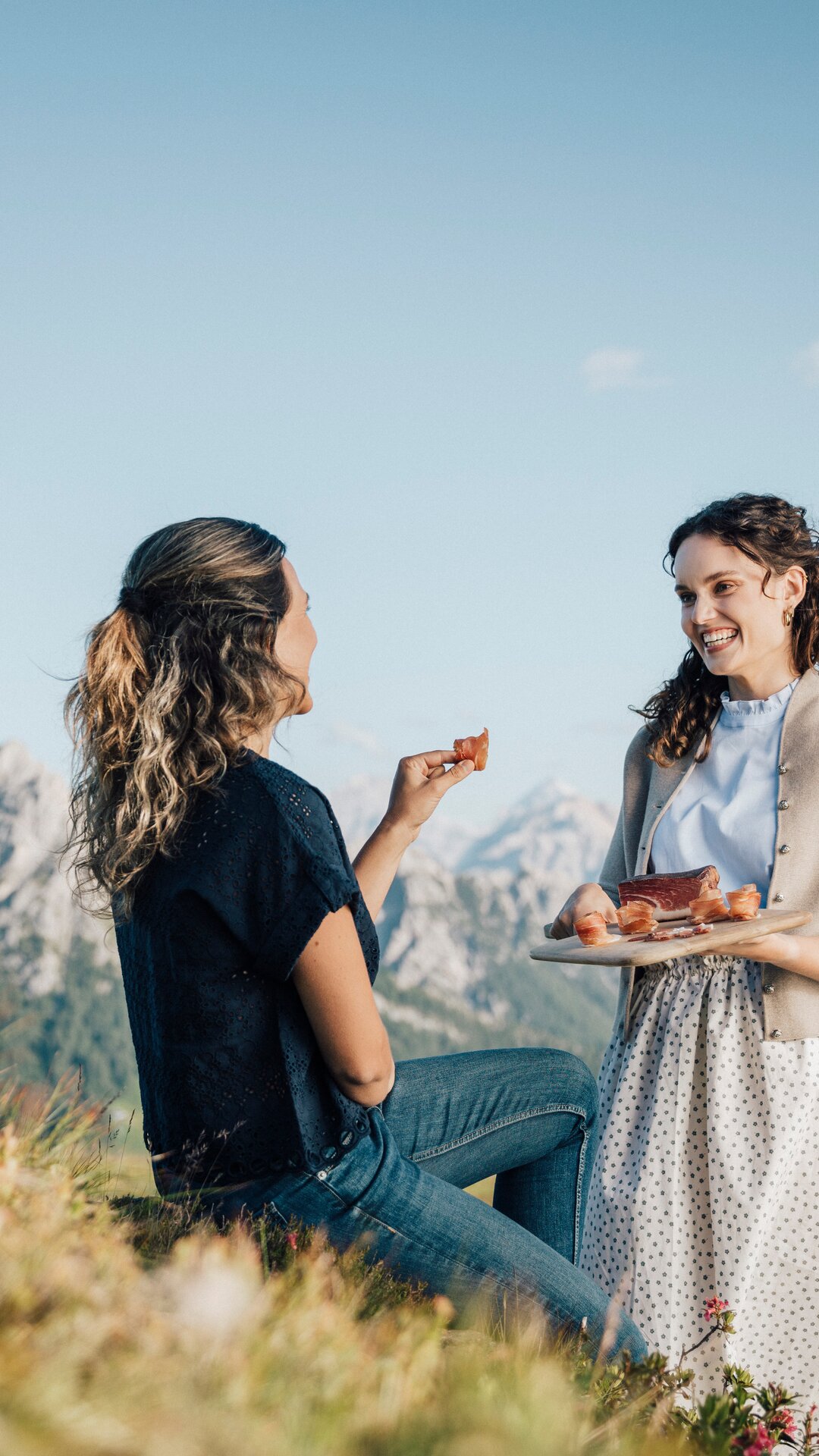 Vor dem Bergpanorama sieht man eine stehende Frau mit einem Brett Speck in den Händen und sie lächelt eine sitzende Frau an, welche sich an dem Speck bedient. | © IDM Südtirol