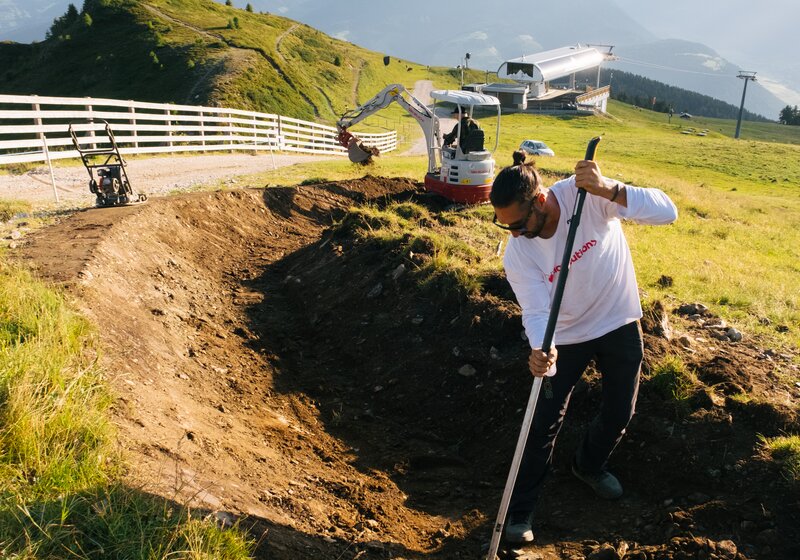 Eine Person bearbeitet mit einer Harke einen Erdhügel auf einem Mountainbike-Trail in Hanglage. | © Daniel Niederkofler
