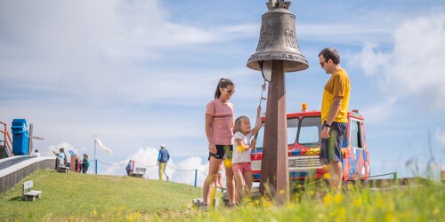 Eine junge Familie steht auf einer Bergwiese, ein kleines Mädchen läutet eine große Glocke.
