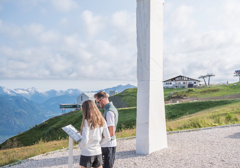 Zwei Personen stehen auf einem Berg neben einer Kunstskulptur und lesen sich das Beschreibungsschild durch, im Hintergrund der leicht bewölkte Himmel.