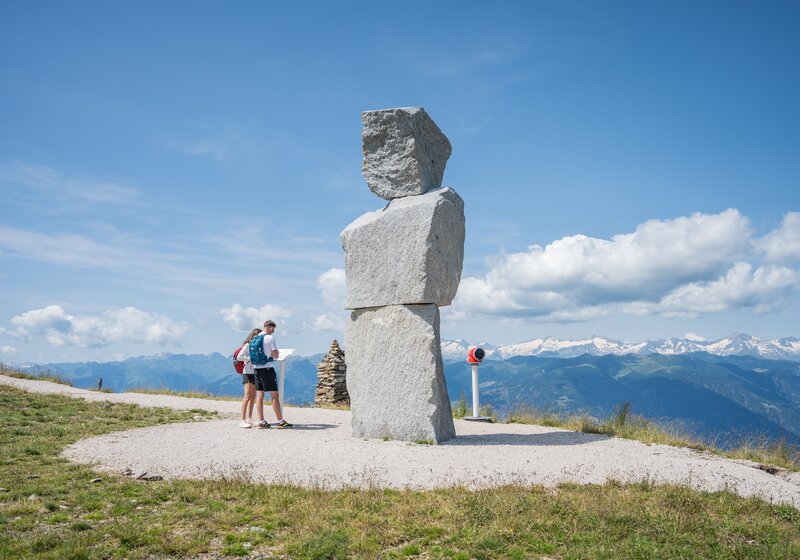 Zwei Personen stehen auf einem Berg vor einer großen Steinskulptur und lesen das Beschreibungsschild, im Hintergrund weitere Berge und der leicht bewölkte Himmel.
