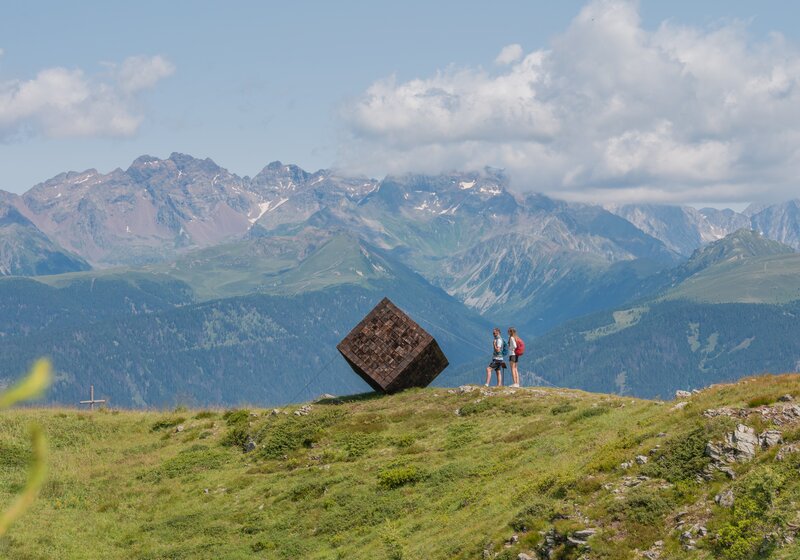 Zwei Personen stehen neben einer riesigen hölzernen würfelförmigen Kunstskulptur auf einem Berg, im Hintergrund weitere Berge.