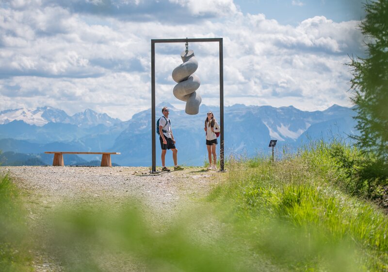 Zwei Personen stehen unter einem hängenden Kunstwerk auf einem Berg, im Hintergrund das Bergpanorama und der leicht bewölkte Himmel.