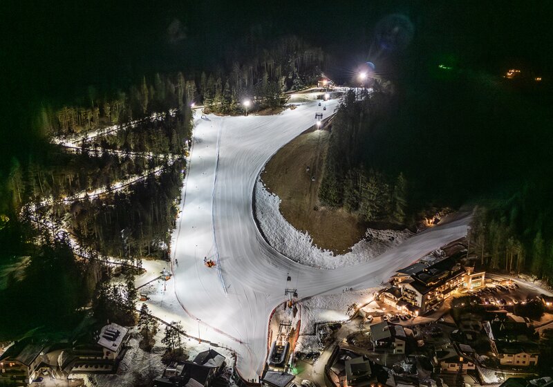 Eine kurvige, beleuchtete Piste schlängelt sich nachts durch einen dunklen Waldhang. | © Gianvito Coco
